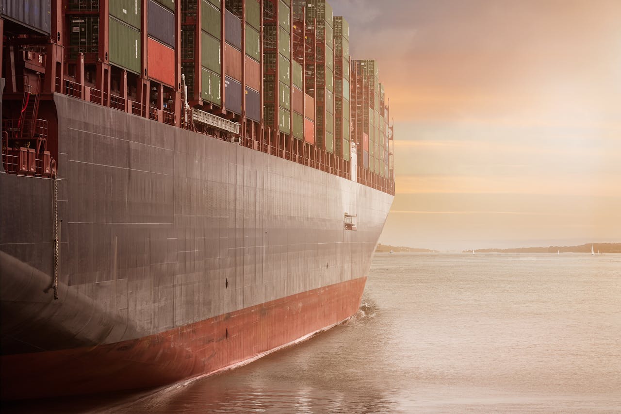 hero-img-01 A colossal cargo ship loaded with containers navigates through calm waters against a vibrant sunset sky.