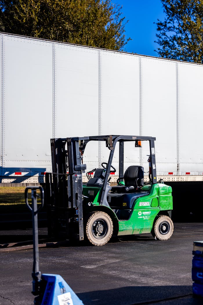 A green forklift parked beside a large white truck outdoors on a clear day.