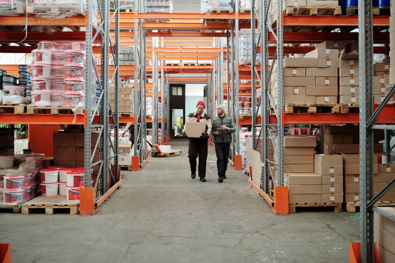 about-01 Two workers handle a package in a spacious warehouse surrounded by shelves stocked with boxes and products.